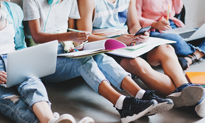 Girl in blue jeans and black sneakers writing lecture in big textbook, sitting on the floor with college friends. Young man typing message on phone while other students working with laptops..